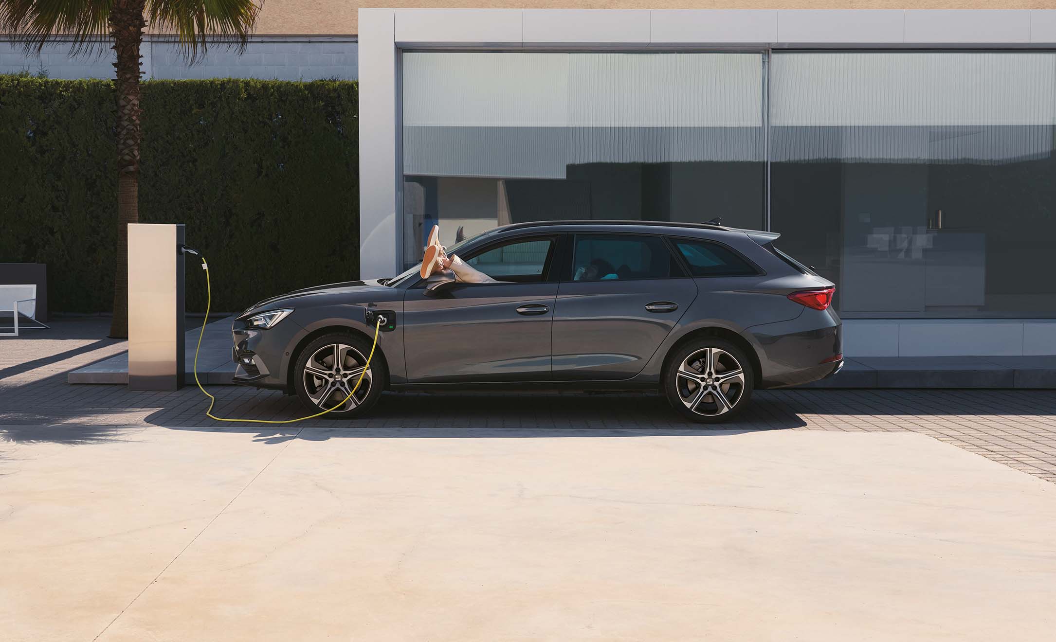 A grey SEAT Leon Estate parked outside a modern house and plugged into a wall charger