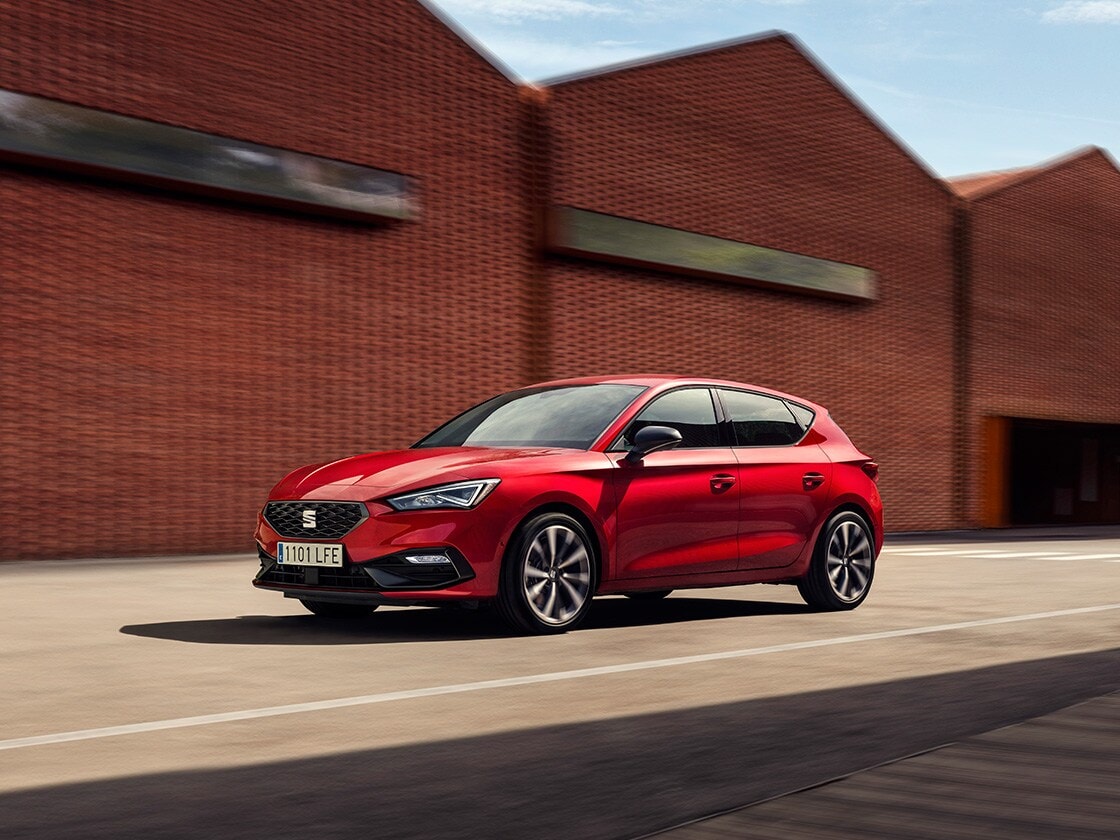 A red SEAT Leon parked under a modern architectural structure. A couple stands nearby, emphasizing the reliability and long-term benefits of SEAT's Service & Maintenance plan for safe and economical vehicle upkeep.