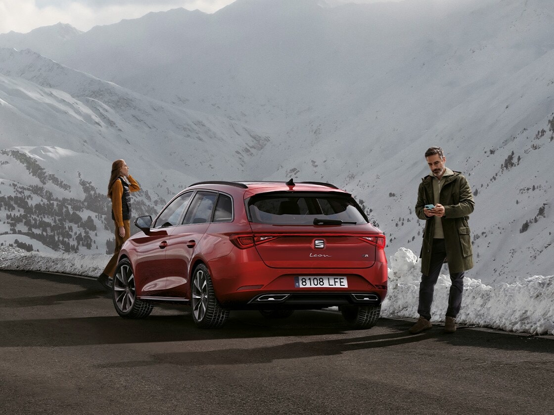 A red SEAT Leon Estate car parked on a mountain road waiting for SEAT Roadside Assistance. Two people standing nearby.