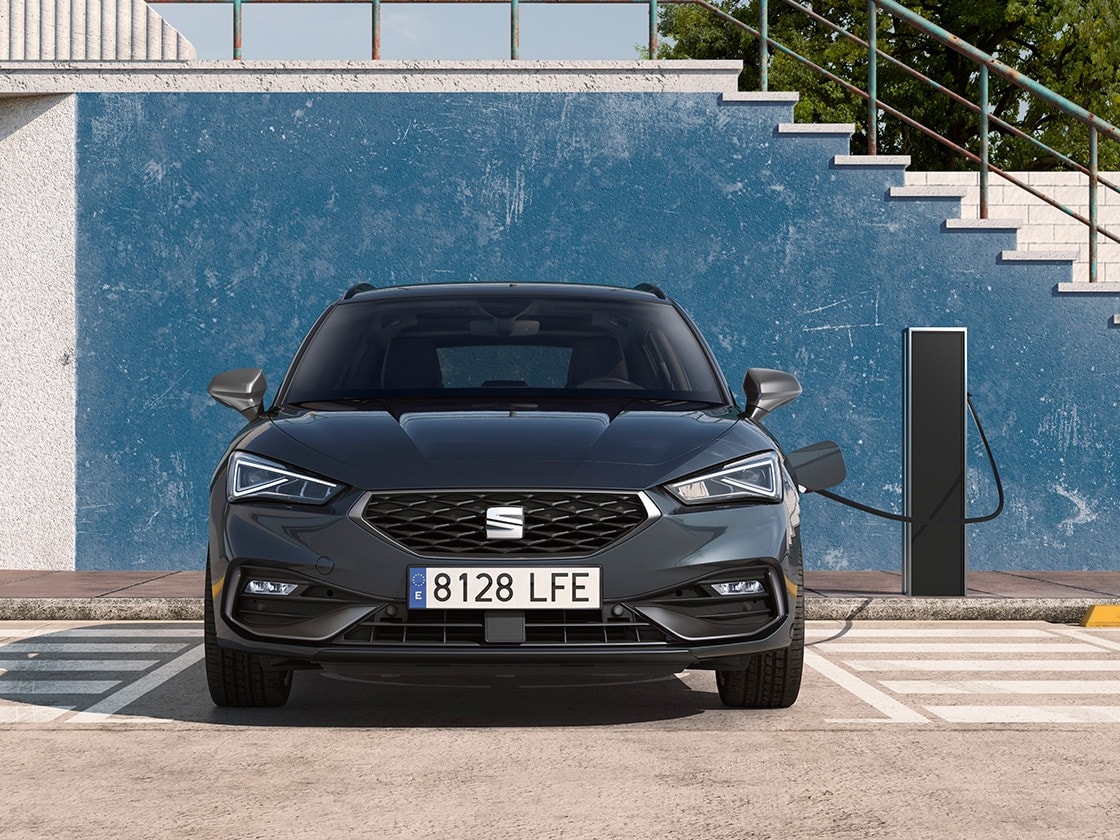 The SEAT Leon Estate e-HYBRID, a hybrid electric car, parked in a parking spot next to a charging station outdoors, showcasing the e manager feature for impressive value.