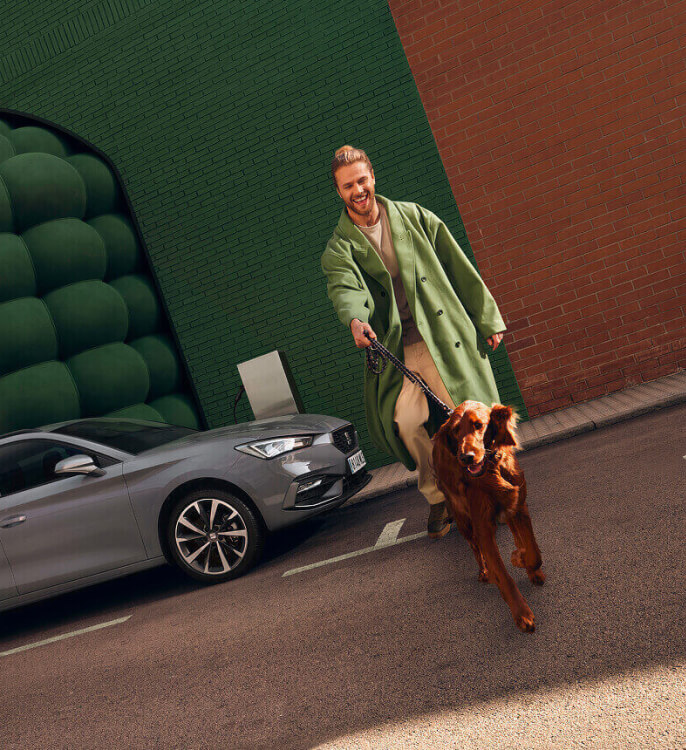 a happy man with a large dog walking away from a grey plug-in hybrid SEAT Leon