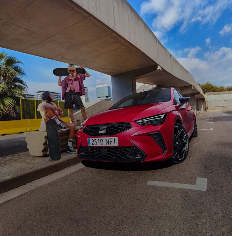 Two people with skateboards sit on a concrete side walk with a new red SEAT Ibiza parked alongside them.