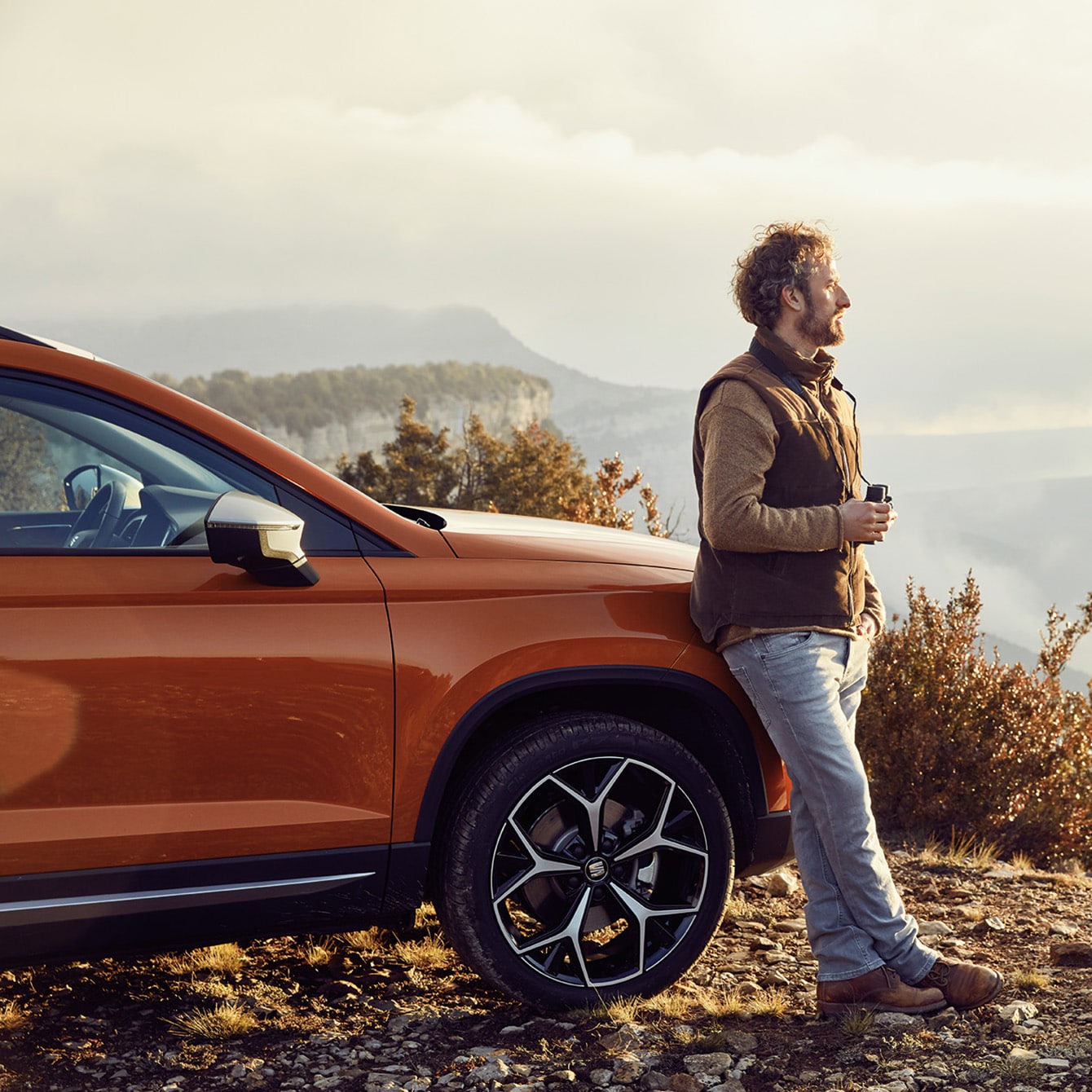 A man leaning against an orange SEAT Ateca on a hill in the countryside and looking off into the distance
