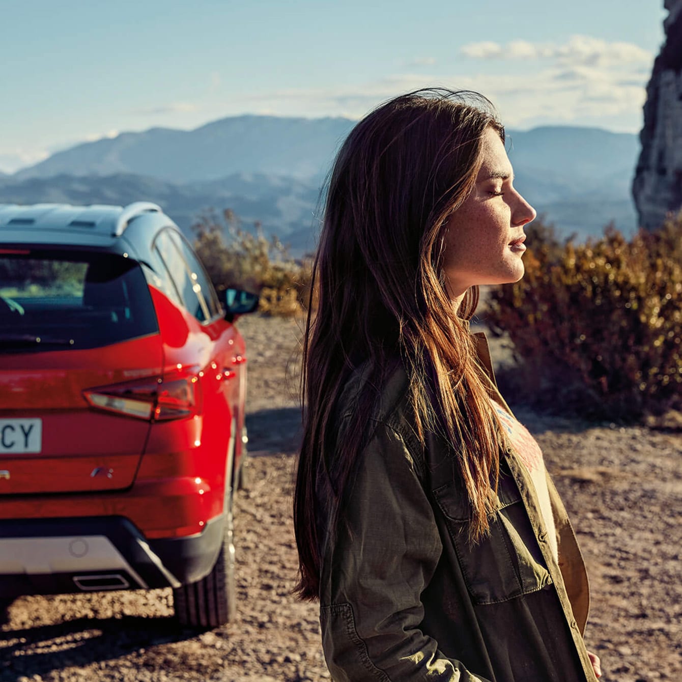 A woman with closed eyes standing next to a red SEAT Arona and facing the sun in a countryside setting with mountains in the background