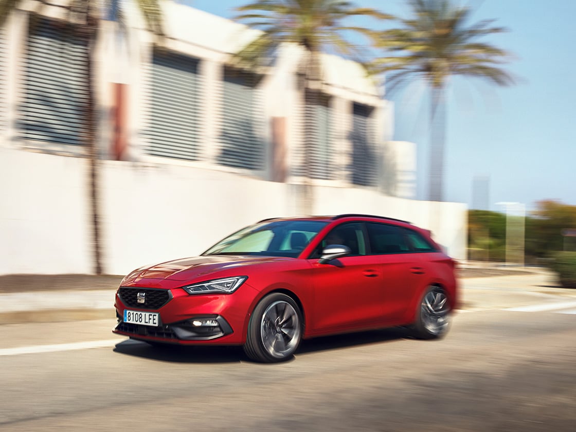 A red SEAT Leon Estate benefits of seat’s service and maintenance plan, in motion in a residential area with a house and palm trees.