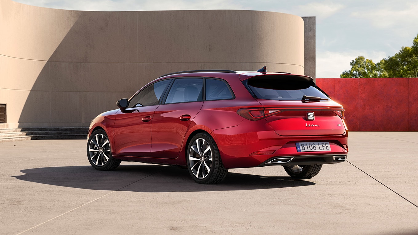 Side rear view of the seat estate car, the SEAT Leon Estate in desire red, parked facing the wall of a building in an outdoors setting. The view showcases the rear design of the car.